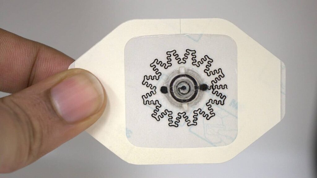 Two fingers are holding an octagonal white patch with a sticker in the middle. The sticker has a circuit design printed on it.