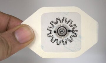 Two fingers are holding an octagonal white patch with a sticker in the middle. The sticker has a circuit design printed on it.